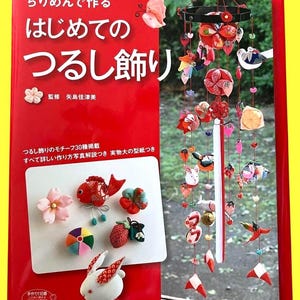 以下が含まれることがあります： 赤い本と、色とりどりの布製オーナメントの吊り下げ式モビール。本の表紙には「初めてのつるし飾り」というタイトルが書かれています。モビールには、花、鳥、幾何学模様など、さまざまな形やデザインが含まれています。