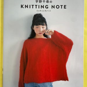 May include: A woman wearing a red knitted sweater and blue jeans. The cover of a book titled "Knitting Note" is visible in the background. The book's title is in English and Japanese.
