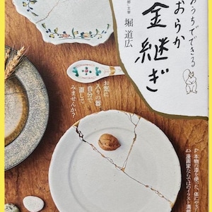 May include: A book cover and several pieces of pottery, including a plate, a spoon, and a bowl, are arranged on a wooden surface. The plate is broken and repaired with gold-colored lacquer, a Japanese art form called Kintsugi. The book title is in Japanese.