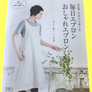 以下が含まれることがあります： 白い縦縞の黒いストライプの白いエプロンを着た女性が、緑の入った花瓶と「Everyday Apron」というタイトルの本のあるテーブルのそばに立っています。本の表紙には日本語のテキストがあります。