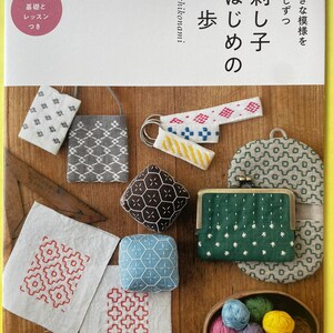 以下が含まれることがあります： 「刺し子はじめの一歩」というタイトルの本。表紙には、刺し子刺繍が施された小さなポーチ、キーホルダー、金属製の口金が付いた緑色の小銭入れ、幾何学模様の布地など、様々な刺し子作品が配置されています。