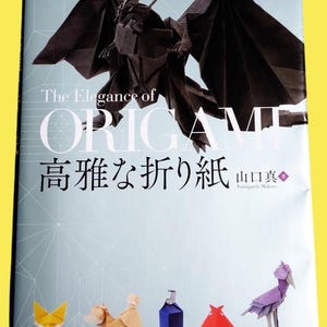 Könnte beinhalten: Buchcover mit dem Titel "The Elegance of Origami" mit einem schwarzen Origami-Drachen. Das Cover zeigt auch japanischen Text und Origami-Tierfiguren, darunter ein Fuchs, ein Hund, eine Flasche und ein Vogel.