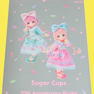 Op de afbeelding: Een magazine cover met de titel "Dollybird vol.32" in roze letters. De cover toont twee Blythe poppen in pastelkleurige jurken en accessoires. De tekst "Sugar Cups" en "20th Anniversary Blythe" is ook zichtbaar.