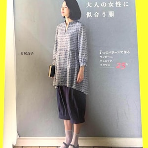 Op de afbeelding: Een vrouw in een tuniek met patroon en marineblauwe culottes staat naast een boek. De boekcover is zwart. De Japanse tekst op de boekcover luidt "Kleding die volwassen vrouwen past".