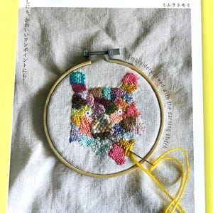 Puede incluir: Un proyecto de bordado colorido en un aro de madera, con un diseño de gato cosido. El texto "Embroidery sewed with the darning stitch" es visible. La portada del libro en el fondo tiene texto japonés.