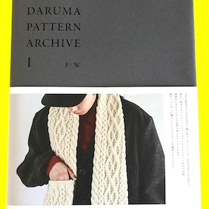 Puede incluir: Un libro de tapa blanda titulado DARUMA PATTERN ARCHIVE 1 F/W con una cubierta gris oscuro y una sección inferior blanca. La portada presenta una fotografía de una persona que lleva una chaqueta negra y una bufanda de punto trenzado color crema.