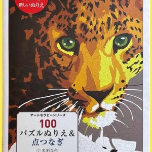 Può includere: Un libro da colorare con un'illustrazione dettagliata del volto di un leopardo. L'immagine è composta da varie sfumature di marrone, giallo e nero, con occhi verdi. La copertina del libro include testo giapponese e le parole "100 Puzzle da colorare & Unisci i puntini".