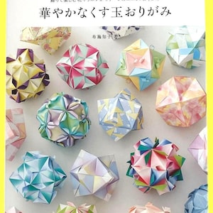 以下が含まれることがあります： 様々な色の折り紙で作られた、幾何学模様のくす玉のコレクション。ピンク、青、緑、黄色など、様々な色の紙球があります。本の表紙には「飾って楽しむ花やリボンがモチーフのユニットおりがみ」と書かれています。