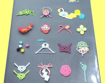 May include: A gray book titled in Japanese with various colorful decorative knots and designs. The knots are in shades of green, pink, red, yellow, and white. The book is on a yellow background.