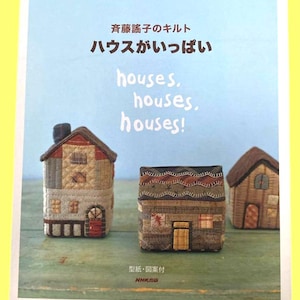 以下が含まれることがあります： 布製のキルトで作られた3つの小さな家の模型が並ぶ本の表紙。テキストには「斉藤謠子のキルト ハウスがいっぱい houses, houses, houses!」と書かれています。背景は風化した質感のライトブルーの表面です。この本には型紙と図案が含まれています。