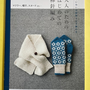 以下が含まれることがあります： 白いスカーフと青いミトンが付いた編み物の本。本の表紙には日本語のテキストがあります。スカーフは折りたたまれ、茶色のボタンが付いています。ミトンは白と青の幾何学模様です。