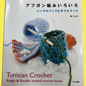 以下が含まれることがあります： 「Tunisian Crochet」というタイトルの本の表紙。青、オレンジ、緑の糸を使ったかぎ針編みの作品例が掲載されています。表紙には日本語のテキストも含まれています。かぎ針編みのプロジェクトは、白い雪の表面に表示されています。
