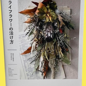 以下が含まれることがあります： 茶色、緑、白の色合いのドライフラワーアレンジメントが特徴の書籍の表紙。 書籍のタイトルは「Dried Flowers」で、上部に日本語のテキストがあります。 表紙には、ハーバリウム、リース、ボタニカルキャンドルに関するテキストも含まれています。