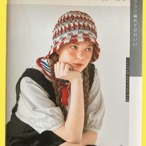 May include: A crochet hat book cover featuring a young person wearing a handmade hat. The hat is red, white, and gray, with a patterned design. The book title is in Japanese, with additional text in Japanese on the right side of the cover.