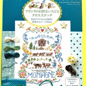 以下が含まれることがあります： フランスの自然をテーマにしたクロスステッチのデザインが特徴の書籍の表紙。山、牛、花などの要素が描かれています。表紙はティール色で、金色の星と日本語のテキストが配置されています。