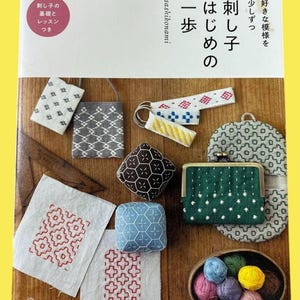 Könnte beinhalten: Eine Sammlung handgefertigter Textilartikel, darunter kleine Taschen und bestickte Stoffquadrate, angeordnet um ein Buch mit dem Titel "Sashiko". Die Artikel weisen geometrische Muster in verschiedenen Farben auf, mit einer Holzschale mit farbigem Garn am unteren Rand.