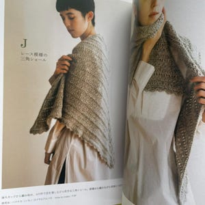 May include: A beige triangular crochet shawl featuring a delicate lace pattern with a scalloped edge. The shawl is displayed in a book, shown draped over the shoulders and wrapped around the neck, showcasing the textured yarn and openwork design.