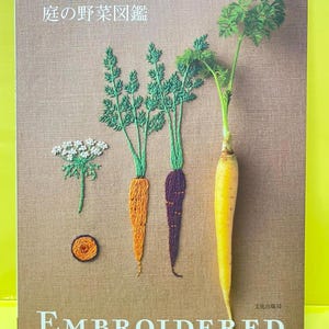 Peut inclure: Une couverture de livre avec un fond texturé marron présentant des illustrations brodées d'une fleur blanche, d'une coupe transversale de carotte et de deux carottes aux fanes vertes. Une carotte jaune est placée à droite. Texte : 青木和子の刺しゅう 庭の野菜図鑑 et EMBROIDERED KITCHEN GARDEN.