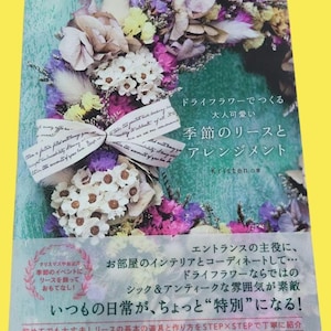 Op de afbeelding: Boekomslag met een bloemenkrans van gedroogde bloemen in wit, paars en geel. De titel is in het Japans, met Engelse tekst op een lint. Het boek gaat over het maken van seizoenskransen en arrangementen.
