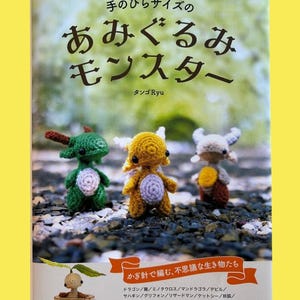 Puede incluir: Una portada de libro en japonés con el título "Amigurumi Monsters", que presenta tres pequeñas figuras de monstruos de ganchillo en verde, amarillo y blanco. El libro también tiene texto en japonés y una pancarta con más texto.
