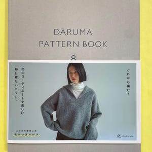 以下が含まれることがあります： DARUMA PATTERN BOOK 8と書かれたグレーの表紙の本。白い帯には、白いタートルネックの上にグレーの襟付きニットセーターを着た人物の写真が印刷されています。帯には日本語のテキストとDARUMAのロゴが記載されています。