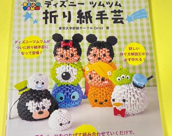 May include: A book cover featuring a colourful illustration of Disney Tsum Tsum characters made from origami. The book title is "Disney Tsum Tsum Origami Crafts" and the subtitle is "Tokyo University Origami Circle Orist".