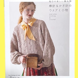 以下が含まれることがあります： ベージュのかぎ針編みのセーターと黄色のスカーフを着た女性が、茶色のセラミック容器を持っています。本の表紙には日本語のテキストと「everyday knitting & crochet」の文字が書かれています。