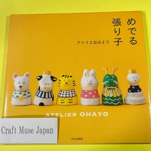 Könnte beinhalten: Ein gelbes Buch mit dem Titel "A  me  de  ru  ri  ko" in Japanisch. Das Buch zeigt Illustrationen verschiedener Tierfiguren, darunter eine Maus, eine Kuh, ein Tiger, ein Kaninchen, ein Drache und eine Figur mit einer Krone. Das Buch trägt den Titel "Atelier Ohayo" und hat den Text "Craft Muse Japan" auf einem weißen Etikett.
