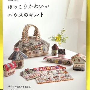 以下が含まれることがあります： 手作りのキルトアイテムのコレクション。トートバッグ、小さな家、ペンケース、プレースマットなど。家をモチーフにしたパッチワークデザインで、様々な色を使用。本の表紙には「ハウスキルト」と書かれています。