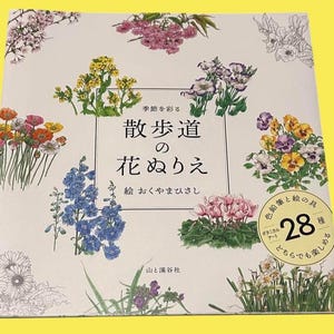 以下が含まれることがあります： クリーム色の表紙に、ピンク、紫、黄色、青色の様々な花のイラストが描かれた正方形の本。日本語のテキストには「花の塗り絵」というタイトルと数字の「28」が含まれています。