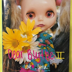 以下が含まれることがあります： 大きな目とブロンドのツインテールを持つ人形が、黄色と白の花を持っている本の表紙。「Dear Blythe II」というタイトルはピンク色の筆記体です。この本は「Scrap Book of Memories」です。