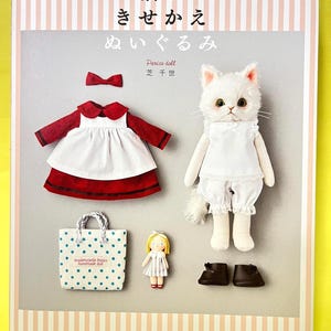 以下が含まれることがあります： Dress-up stuffed Catと題された手芸本の表紙。白い猫のぬいぐるみ、赤いドレスと白いエプロン、小さな人形、青い水玉模様のトートバッグ、赤いヘアリボン、ダークブラウンの靴がライトグレーの背景に配置されています。