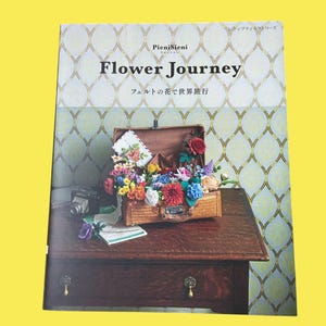 以下が含まれることがあります： 「Flower Journey」というタイトルの本（日本語テキスト付き）には、カラフルなフェルトの花でいっぱいのスーツケースが描かれています。ヴィンテージカメラと地図が、模様のある壁紙の背景を持つ、濃い木製のテーブルの上にあります。