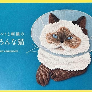 以下が含まれることがあります： 青い目の茶色と白の毛並み、装飾的な襟の猫の刺繍が施された本の表紙。表紙のテキストは「フェルトと刺繍の猫」と「ahoy embroidery」で、背景はティール色、縁は黄色です。