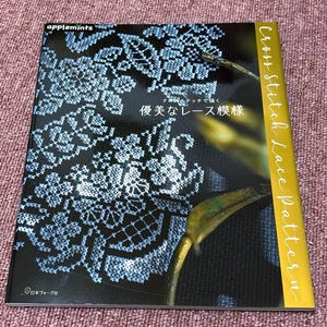 Op de afbeelding: Een boek getiteld "Cross Stitch Lace Pattern" met een zwarte kaft met een wit en blauw kanten ontwerp. Het boek heeft het "applemints" logo en Japanse tekst. De rug is goudkleurig.