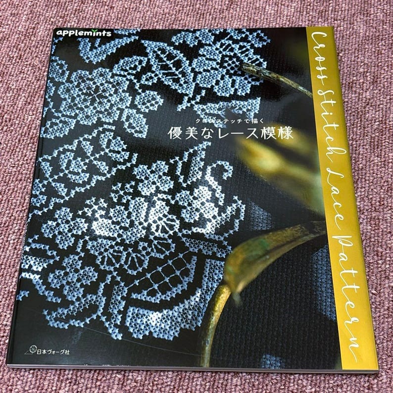 Op de afbeelding: Een boek getiteld "Cross Stitch Lace Pattern" met een zwarte kaft met een wit en blauw kanten ontwerp. Het boek heeft het "applemints" logo en Japanse tekst. De rug is goudkleurig.