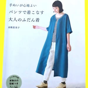 Könnte beinhalten: Eine Frau trägt einen langen, teal-farbenen Mantel über einem gestreiften Hemd und einer weißen Hose. Das Bild stammt aus einem japanischen Nähbuch mit japanischem Text. Das Buchcover ist sichtbar.