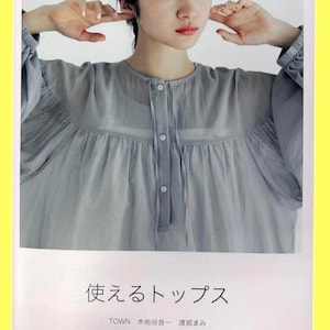 May include: A light gray long-sleeved blouse with a round neckline, button-down front, and gathered fabric details. The book cover features the Japanese text "使えるトップス", "TOWN", "木地谷良一 渡部まみ", "サイズ1・2の縫い代つき実物大パターン", and "文化出版局".