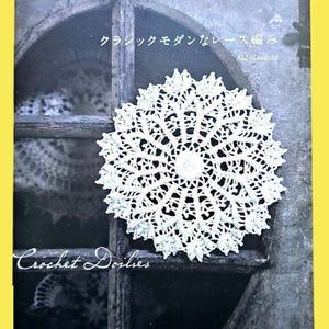Può includere: Copertina di un libro con un centrino all'uncinetto bianco su sfondo scuro. Il titolo del libro è "Crochet Doilies", con testo giapponese sopra il centrino. Il centrino è circolare con intricati motivi in pizzo.