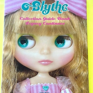 以下が含まれることがあります： 表紙に人形の顔が描かれたBlytheコレクションガイドブック。人形は大きなターコイズ色の目、金髪、ピンクのリボンをしています。本のタイトルはティールとピンクです。