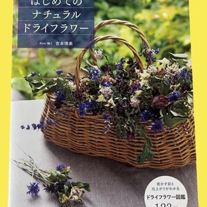 May include: A woven wicker basket filled with dried flowers in shades of purple, blue, and white. The basket has two arched handles and sits on a light gray surface. A book with Japanese text is in the background.