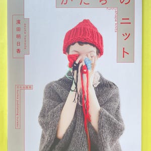 May include: A book cover featuring a person wearing a bright red knit beanie and a chunky grey knit sweater. The person holds several strands of colourful yarn in front of their face. The text reads "かたちのニット", "funny shape knits", "asuka hamada", and "文化出版局 bunka publishing bureau".