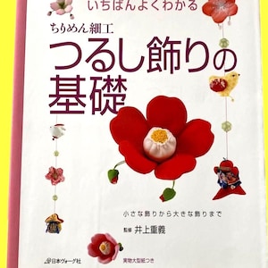 May include: A book cover featuring Japanese text and decorative hanging ornaments. The cover has a large red flower with a white center, and smaller hanging ornaments. The title is in large Japanese characters.