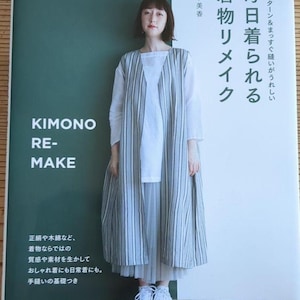 以下が含まれることがあります： 「KIMONO RE-MAKE」のタイトルと日本語のテキストが書かれた本の表紙の前で女性が立っています。彼女は白いトップスと、長いストライプの着物風の衣服を着ています。本の表紙にはテキストがあり、緑と白の背景があります。