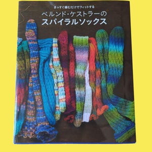 Könnte beinhalten: Ein Buchcover mit japanischem Text und einer Präsentation von bunten, gestrickten Spiralsocken. Die Socken sind in verschiedenen Farben, darunter Blau, Grün, Rot und Lila. Die Socken werden vor einem dunkelbraunen Hintergrund präsentiert.
