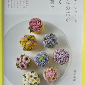 以下が含まれることがあります： 「白あんでつくる あんの花が咲くお菓子」というタイトルの書籍の表紙。ピンク、黄色、紫、青、白の繊細なあんフラワーで飾られた7つのカップケーキが写っています。テキストには、あんことクリームチーズを使ったフラワーデコレーションの詳細が記載されています。