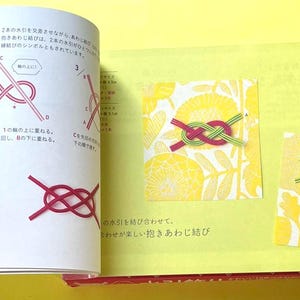 Puede incluir: Libro abierto con texto japon&eacute;s e ilustraciones sobre nudos. La p&aacute;gina izquierda muestra diagramas de los pasos para anudar. La p&aacute;gina derecha muestra dos nudos terminados, uno rojo y verde, el otro azul y verde, sobre fondos florales amarillos.