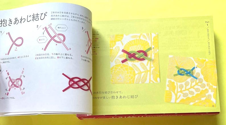 Puede incluir: Libro abierto con texto japon&eacute;s e ilustraciones sobre nudos. La p&aacute;gina izquierda muestra diagramas de los pasos para anudar. La p&aacute;gina derecha muestra dos nudos terminados, uno rojo y verde, el otro azul y verde, sobre fondos florales amarillos.