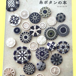 Op de afbeelding: Een boek getiteld "Draadknoppen" met Japanse tekst en diverse handgemaakte draadknoppen in wit, marineblauw en groen. De knopen hebben ingewikkelde geometrische patronen en sterpatronen.