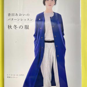 以下が含まれることがあります： 香田あおいのパターンレッスン 秋冬の服というタイトルのソフトカバーの裁縫教本。表紙には、鮮やかなブルーのロングコートを羽織り、ライトグレーのトップスとパンツを着用した人物の写真が掲載されている。本には日本語のテキストが含まれている。